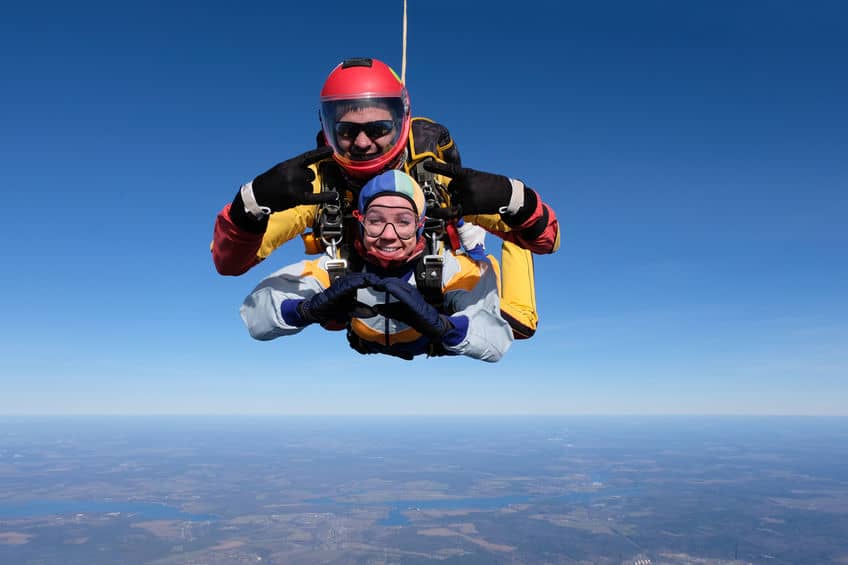A skydiving tandem pair - the skydiver in a red helmet and yellow and black suit and his student in a blue helmet and white suit are in freefall above a landscape of mixed green and brown, under a clear blue sky.