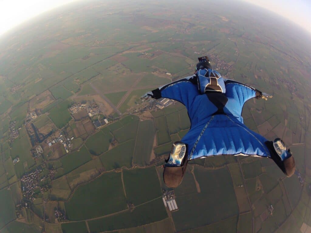 A wingsuit pilot in a blue wingsuit is soaring over an expansive green and brown landscape below and a blue sky above.