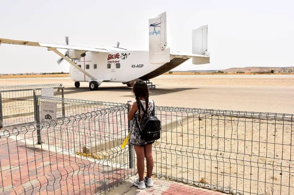 A small girl in a black and white dress wearing a backpack observing a small white propeller plane, parked on a tarmac in a desert landscape.