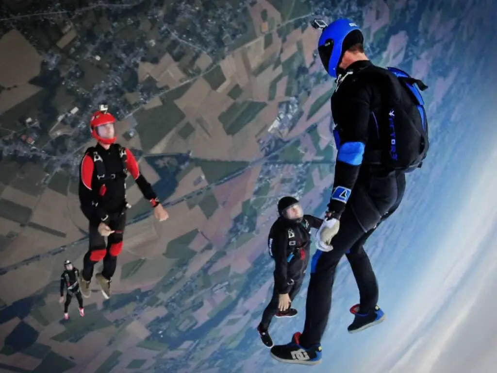 Four skydivers in mid-air, each in a different position, with a view of fields and roads on the ground below.