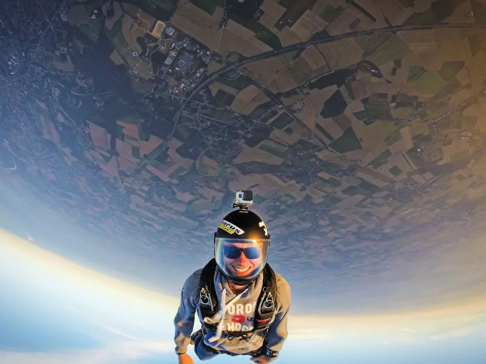Skydiver in blue and black suit freefalling above patchwork of fields and roads. The skydiver is wearing shades underneath the helmet with a big smile on his face.