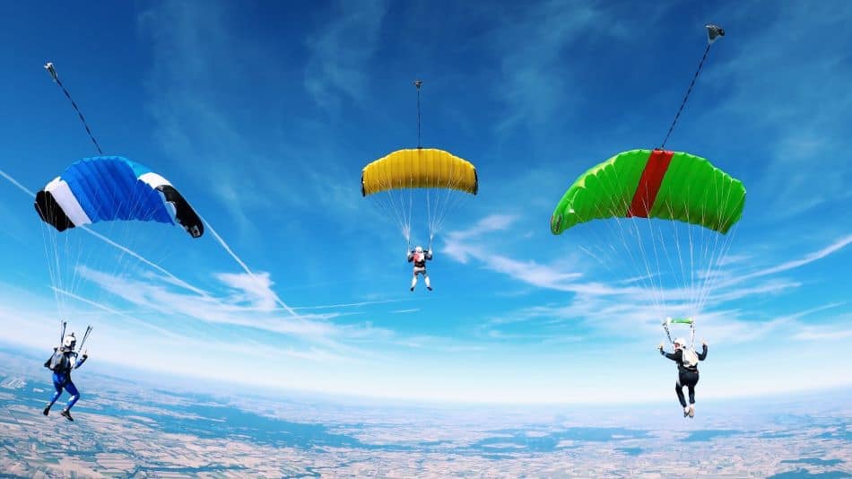 Three skydivers with colorful parachutes are descending towards the ground with a clear blue sky in the background