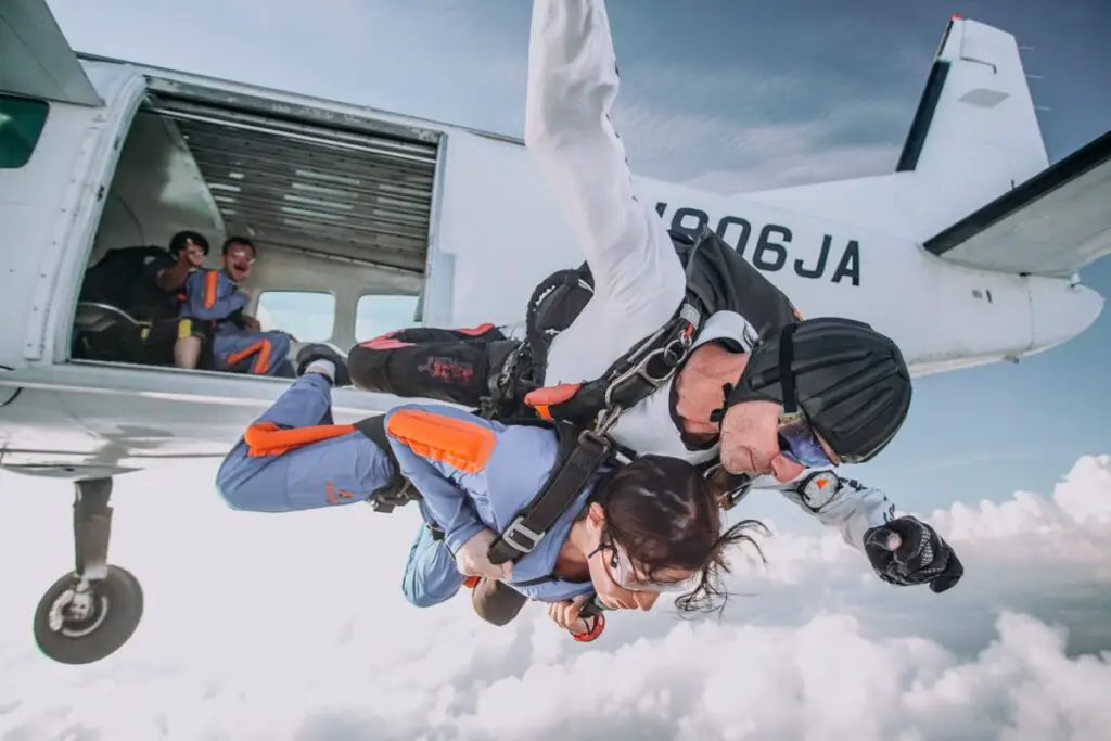 A tandem skydiving pair - the instructor wearing a black helmet and white suit and his lady student in a light blue suit with orange accents in head-down position exiting a small white airplane against a backdrop of a cloudy sky.