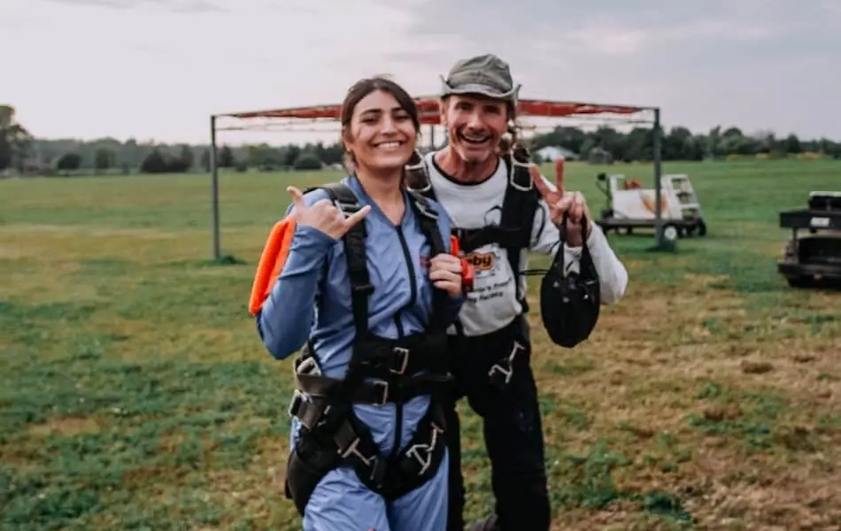 Two skydivers in jumpsuits and harnesses on a field of grass posing in front of the camera. 
