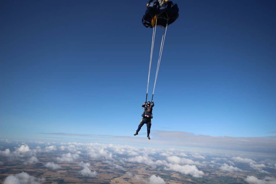 A skydiver in a blue suit and black helmet is in freefall above a landscape of fields and clouds. The parachute was just deployed, and the sky is clear with visible white clouds.
