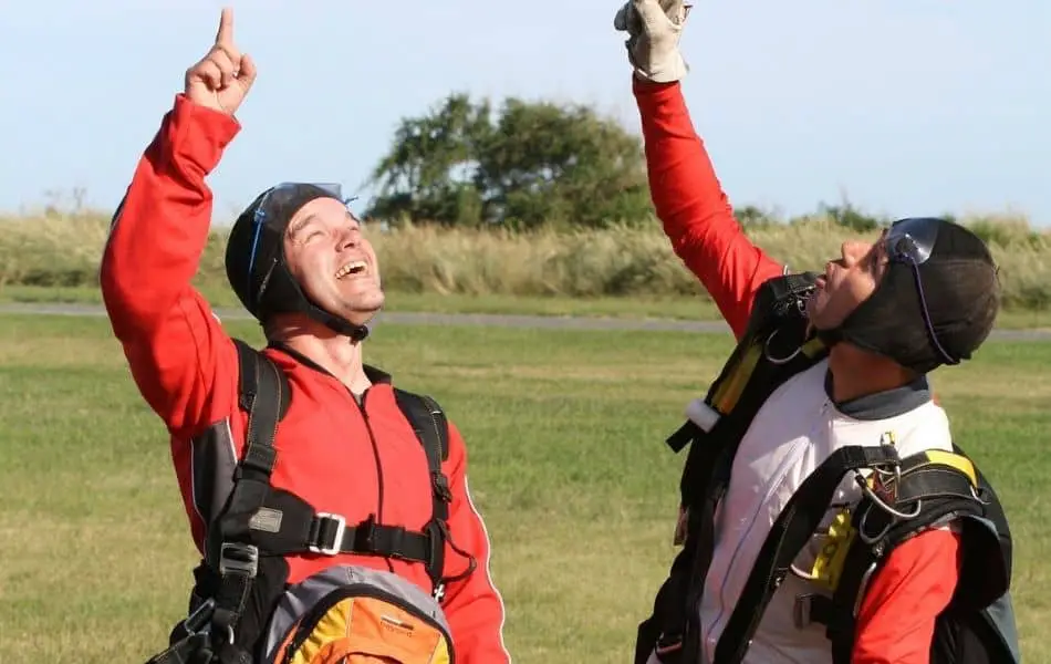 Two skydivers in red and white suits, celebrating a successful jump with arms raised pointing up.