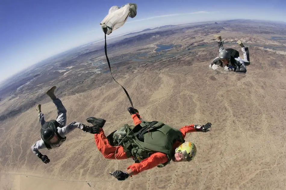 A group of three skydivers in mid-air with one skydiver’s parachute deploying above a desert-like landscape.