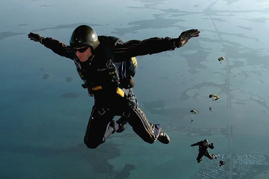 A skydiver in black suit, mid-air with arms spread, along with other skydivers that have deployed their parachutes  above a landscape.