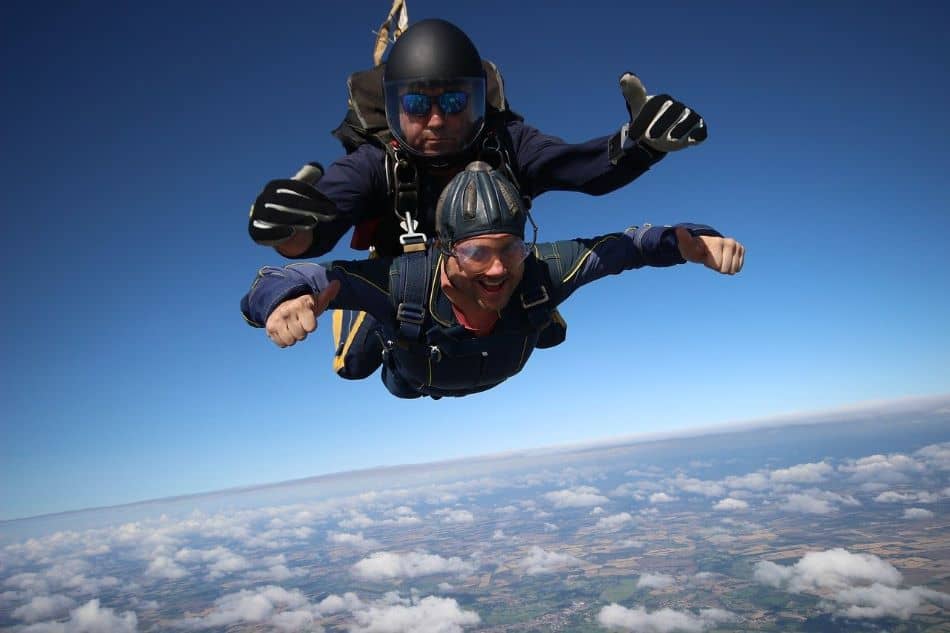 Two people skydiving in tandem, wearing blue and black jumpsuits, above a cloudy sky with a landscape below in green and brown land with mountains and rivers.