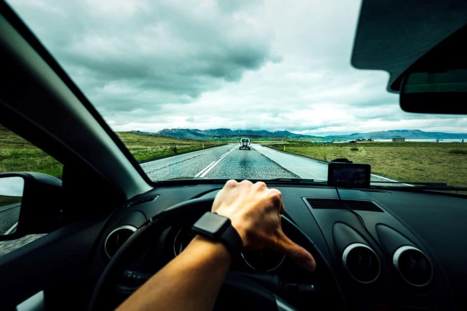 A photo of a person’s hand on the steering wheel of a car, facing the road, driving on a highway with mountains in the foreground and fields of grass on the side.