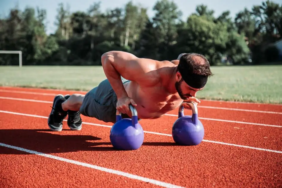 A man doing push-ups on two blue kettlebells on a red track field. He is shirtless, wearing gray shorts, and the background features trees and a clear blue sky.