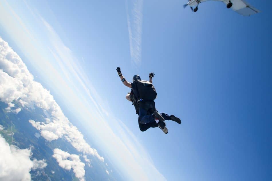 A skydiving tandem pair are captured in mid-air against a backdrop of a clear blue sky scattered with white clouds. They are both dressed in dark attire with helmets for safety, and their bodies are positioned in a classic freefall stance with arms and legs extended. In the top right corner of the image, a small white airplane can be seen, likely the one they jumped from.