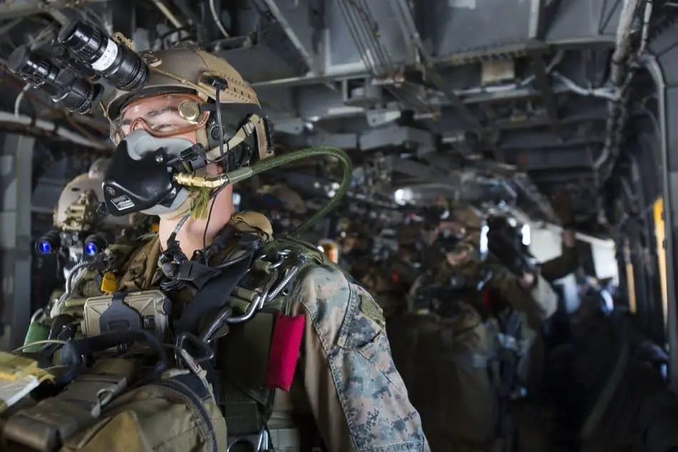 A group of soldiers in full gear are seated inside a military aircraft. They are equipped with helmets featuring night vision goggles, camouflage uniforms, body armor, backpacks, and weapons. The interior of the aircraft shows additional equipment and seating arrangements.