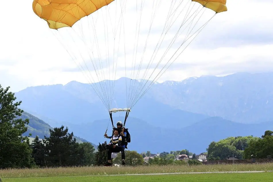 A tandem skydiving pair in blue and black suits gliding over a green field with distant mountains shrouded in haze. The landscape includes trees and houses from afar.