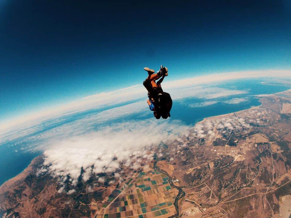A skydiving tandem pair in blue and black jumpsuits are in freefall, positioned head-down above a picturesque landscape featuring a coastline and agricultural fields. The sky is blue with scattered clouds.