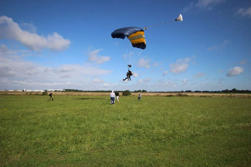 A skydiving tandem pair in black suits are parachuting onto a grassy field under a blue and yellow parachute. The sky is clear with a some clouds, and four onlookers are present on the ground.