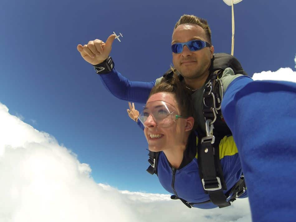 A skydiving tandem pair wearing blue jumpsuits in freefall, set against a backdrop of a blue sky scattered with white clouds. In the upper part is a small image of a white plane, likely the one they jumped out from.