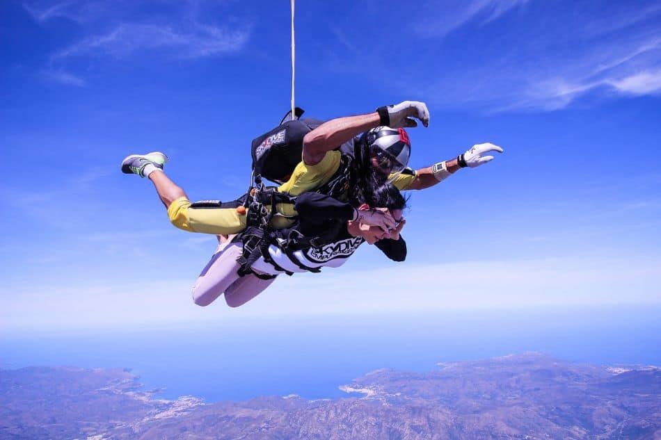 A tandem skydiving pair over a breathtaking view of the blue ocean and coastline. The person in the front is in a black and white jumpsuit, while the one behind is in a yellow jumpsuit. Both are equipped with helmets and goggles for safety. The parachute has just been deployed. 