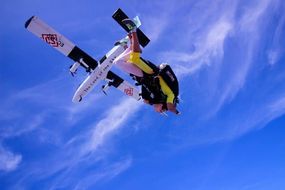 A skydiving tandem pair in yellow jumpsuit and black helmet, captured in an head-down freefall position. In the background, a white and black plane, likely the one they jumped from, set against a bright blue sky adorned with wispy clouds.