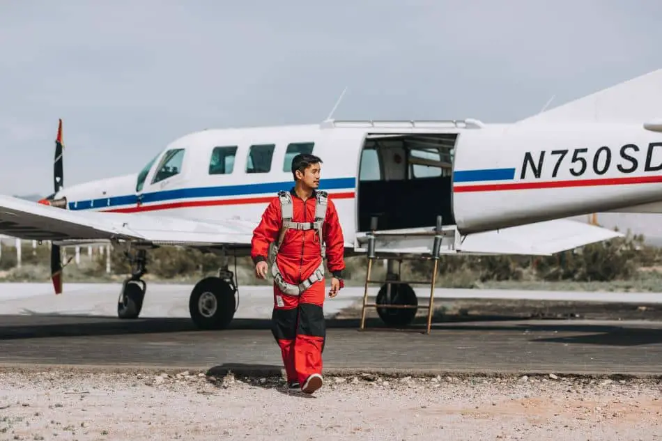 A person in a red jumpsuit is walking in front of a white and blue propeller plane set against a desert landscape and clear blue sky.