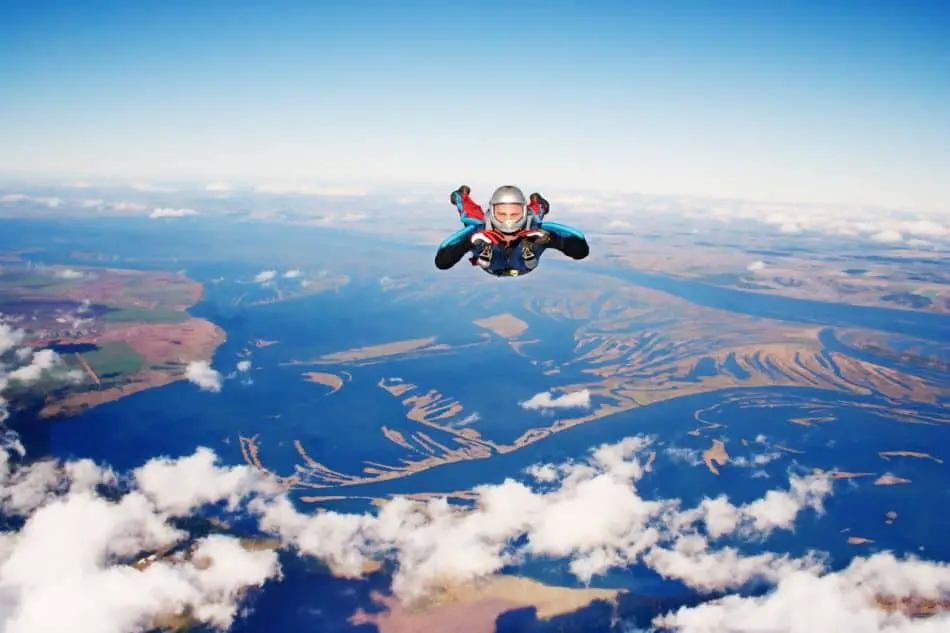 A skydiver in a blue and red suit is in freefall above a landscape of rivers and fields. The skydiver is positioned with arms and legs spread out, against a backdrop of a clear blue sky with white clouds.