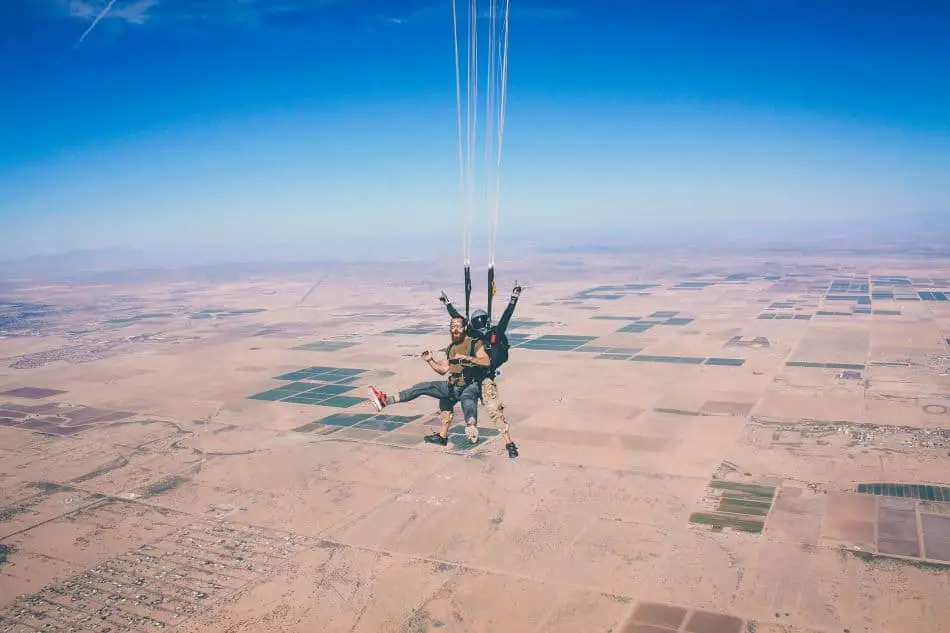 A skydiving tandem pair in mid-air with their parachute open, above a vast landscape of fields and terrains.