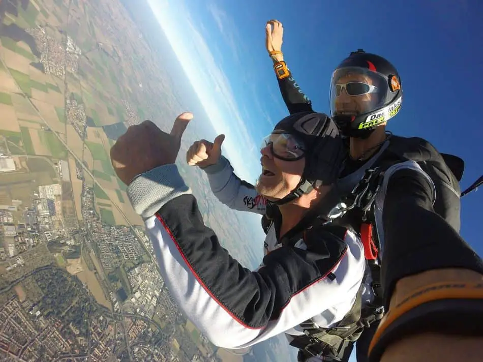 A skydiving taandem pair, with the student in the front giving a thumbs up, and the instructor in the back, hands stretched against the backdrop of a clear sky and landscape below.