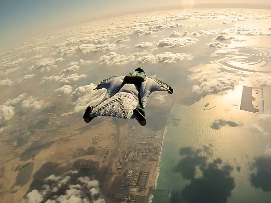 A wingsuit pilot in a black and white wingsuit with clouds everywhere soaring over a city and the shimmering ocean with a sunset behind.