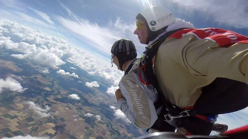 A skydiving tandem pair in red and white jumpsuits and helmets, in freefall above a patchwork of fields and clouds. The student's arms are in the chest and the instructor behind has his arms outstretched, against a backdrop of a vivid blue sky dotted with fluffy white clouds and the earth below, marked by the green and brown hues of the fields.