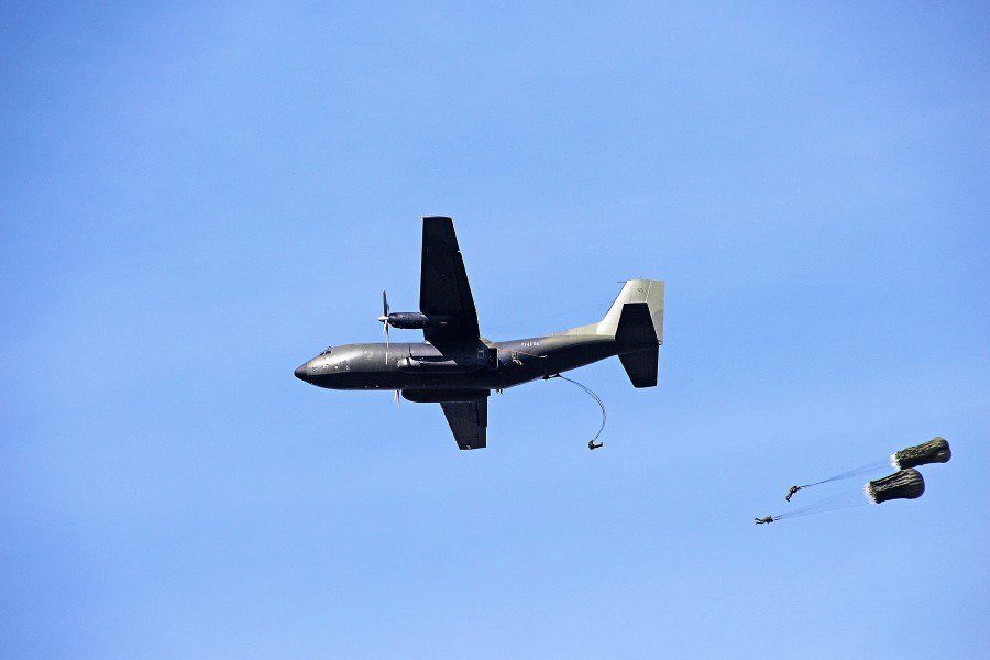 A black plane flying in the sky and three skydivers with one's parachute seemed to be caught buy the plane and the two getting blown away, they are all set against a clear blue sky background.