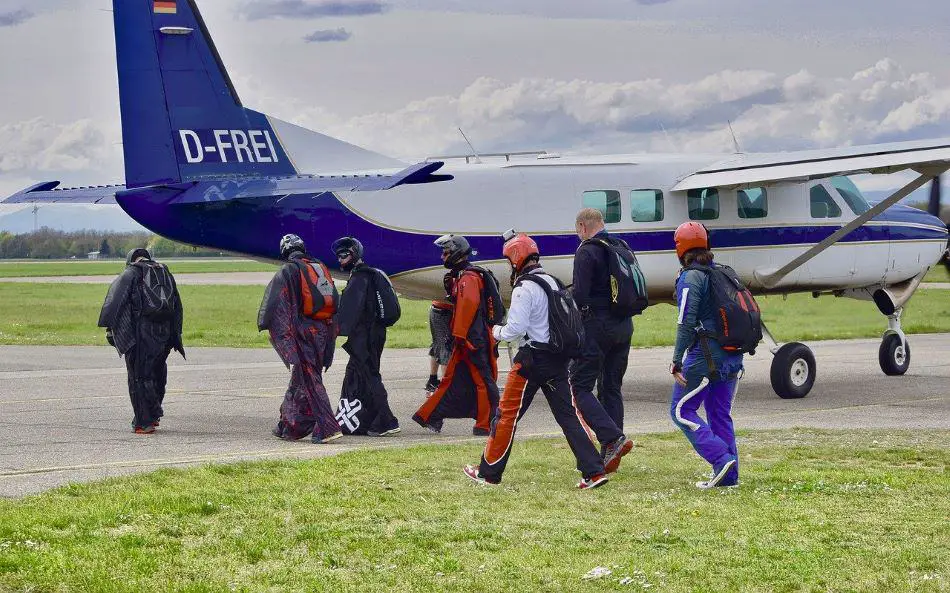 A group of wingsuit pilots in colorful wingsuits walking towards a small airplane on a green airfield under a blue sky and thick white clouds.