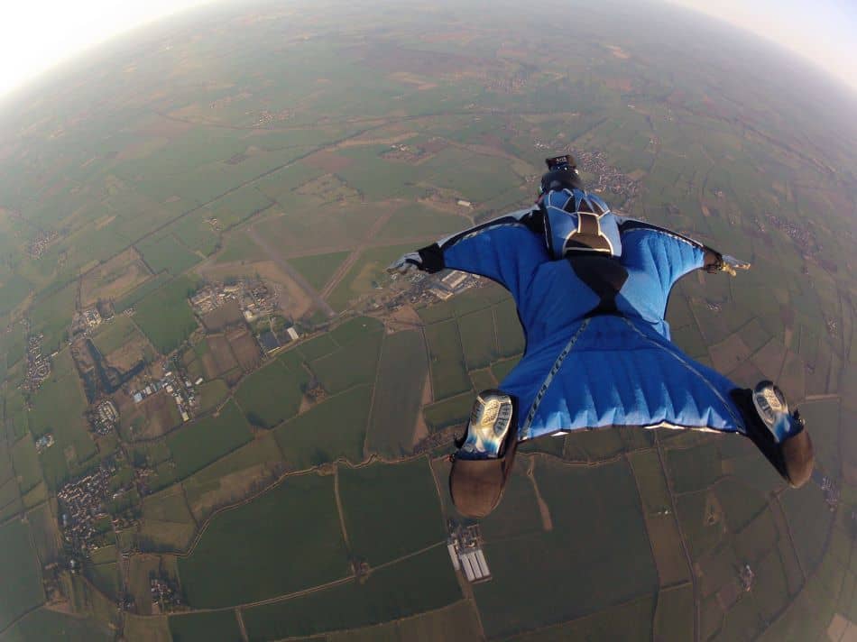 A wingsuit pilot in a blue wingsuit is soaring, captured mid-air with a view of the green landscape below.