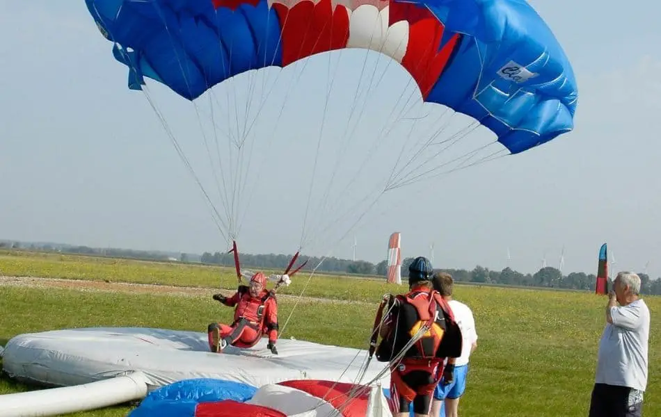 A skydiver in a red jumpsuit landing on an inflatable pad with a blue and red parachute. There are people around, one wearing a red and black jumpsuit and two others in casual attire that seem to be involved in the activity. The background is a grassy field and a blue sky.