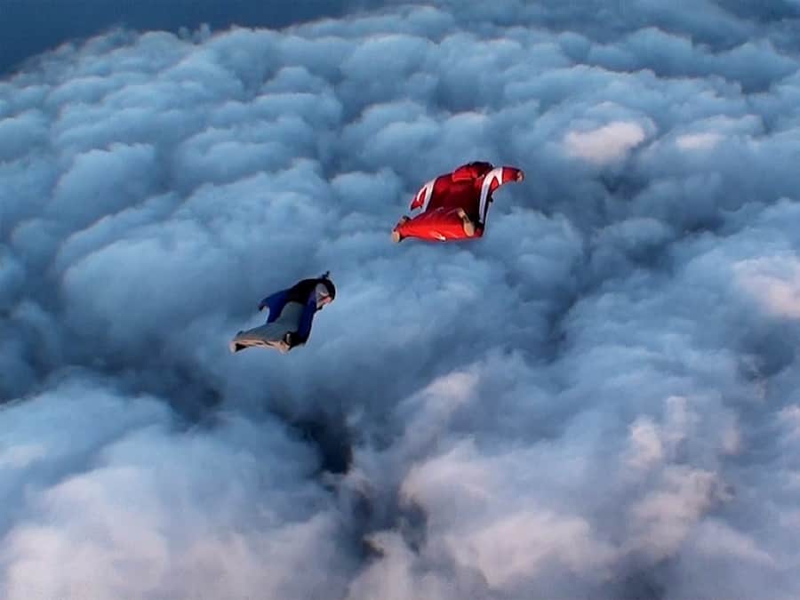 Two wingsuiut pilots in blue and red wingsuts gliding above a sea of clouds.