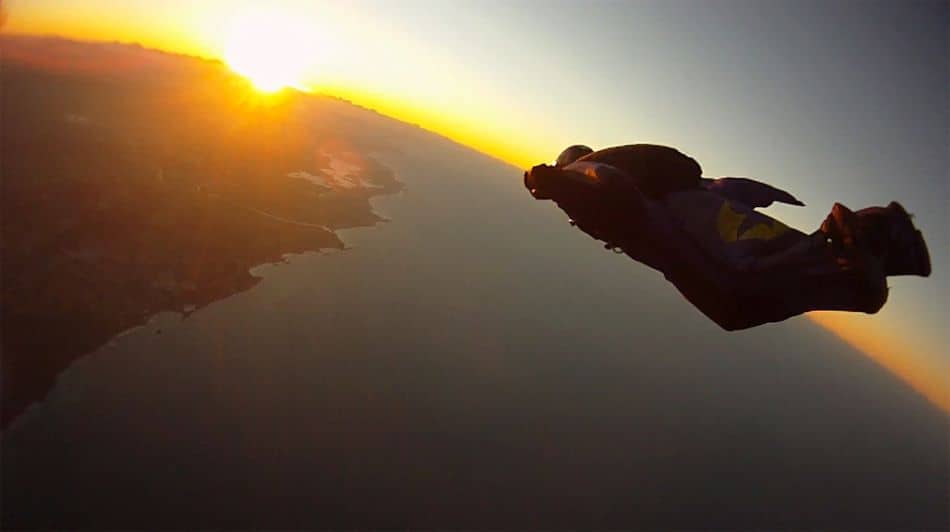 A wingsuit pilot glides in the air against a sunset sky and a vast landscape below.
