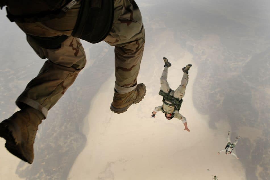 A soldier in camouflage attire is captured mid-air, with three other soldiers and the cloudy atmosphere below them, illustrating an intense moment of freefall during a daytime jump.