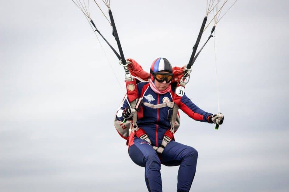 A skydiver in a red and blue suit and helmet controls the parachute lines against a cloudy sky.