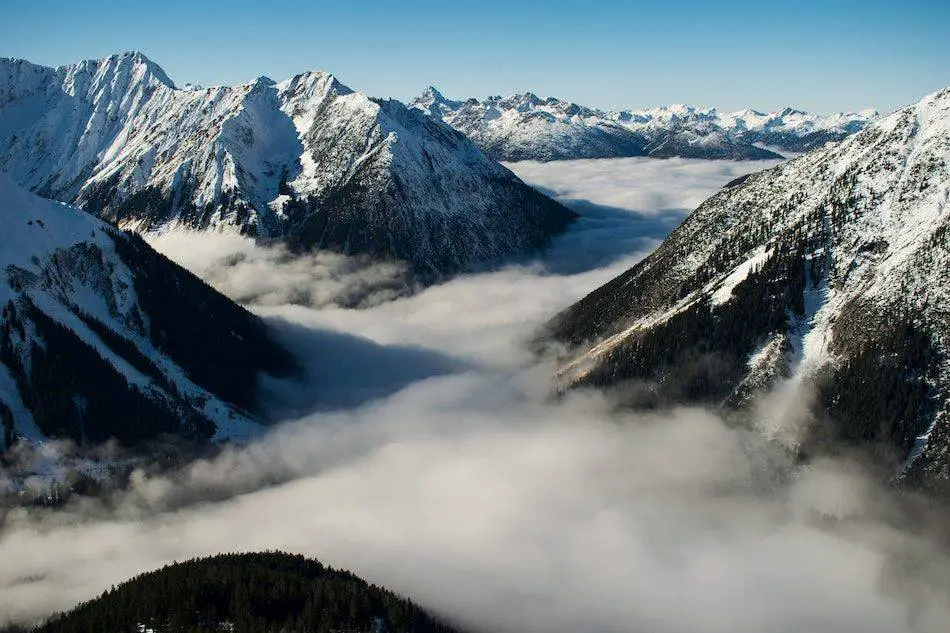 A breathtaking view of snow-capped mountains with valleys filled with thick, flowing clouds under a clear blue sky.