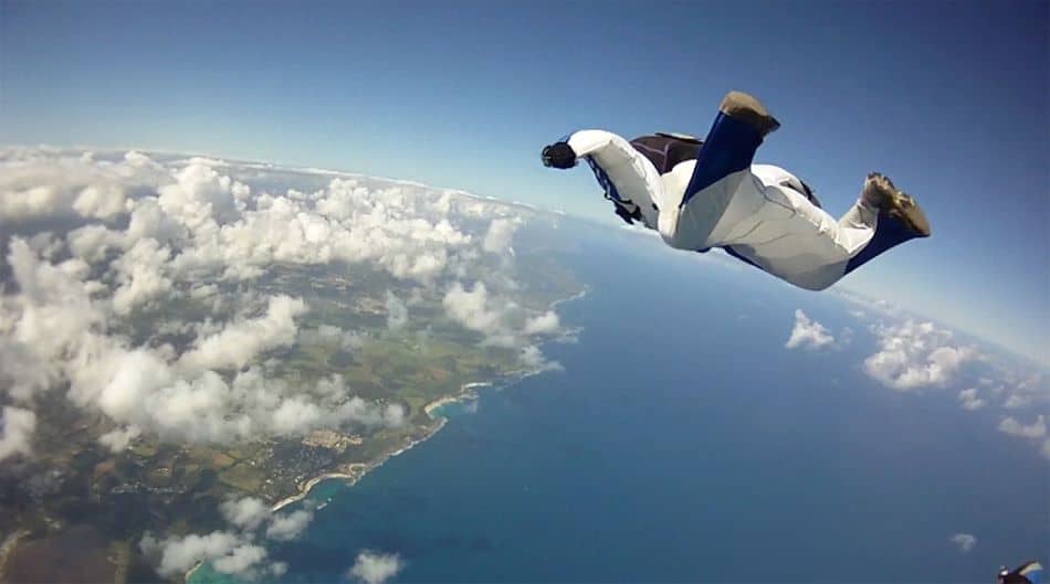 A wingsuit pilot in a white and black suit is captured mid-air against a backdrop of scattered clouds, blue skies, and an expansive view of the coastline below.