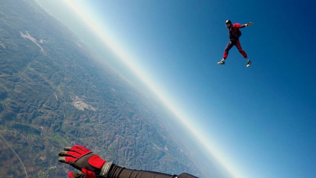 A skydiver in black and red suit freefalls against the backdrop of earth and sky, with another skydiver's hand visible in the foreground.