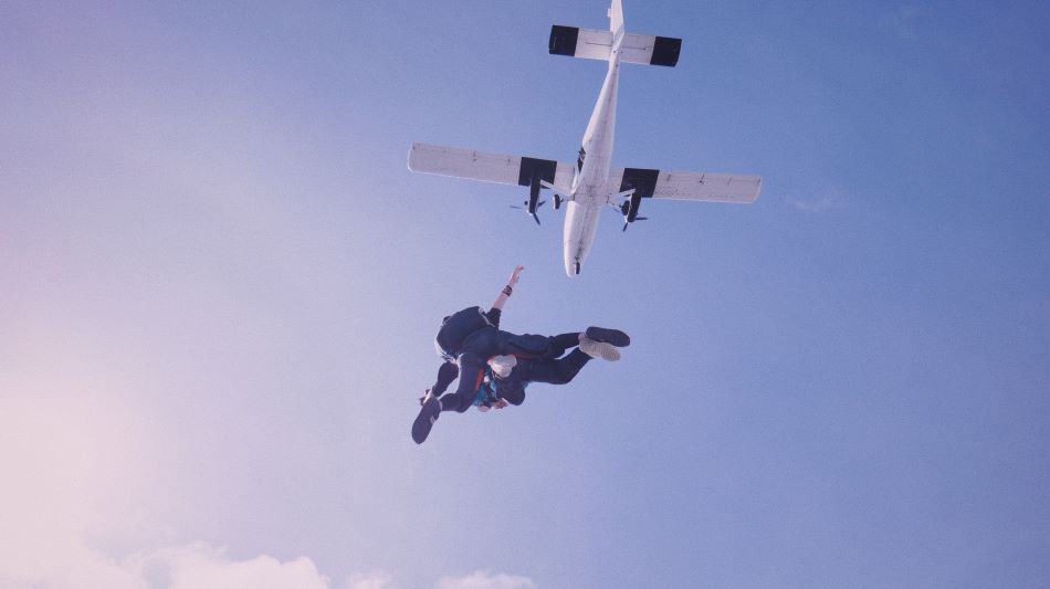 A skydiving tandem pair in mid-air, having just exited a white airplane against a clear blue sky.