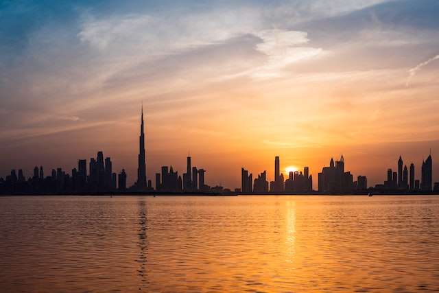 A serene view of a city skyline silhouetted against a golden sunset, with the calm waters of a lake or sea reflecting the warm hues of the sky and buildings.