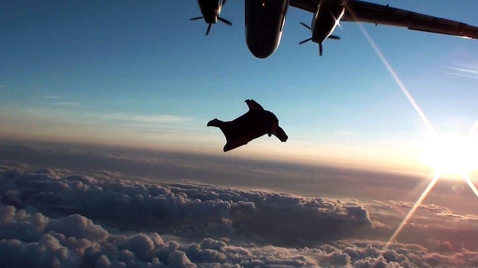 A wingsuit pilot in mid-flight, soaring away from an aircraft above a breathtaking view of clouds illuminated by the setting or rising sun.