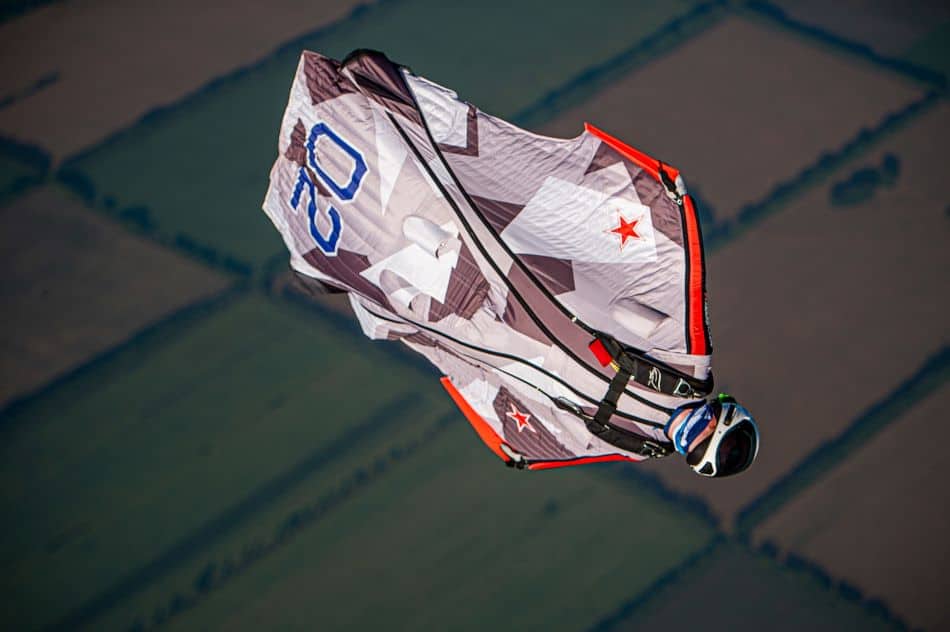 A wingsuit pilot in a white and gray suit with blue numbers 02 and red stars, gliding against the backdrop of a patchwork of green fields below.