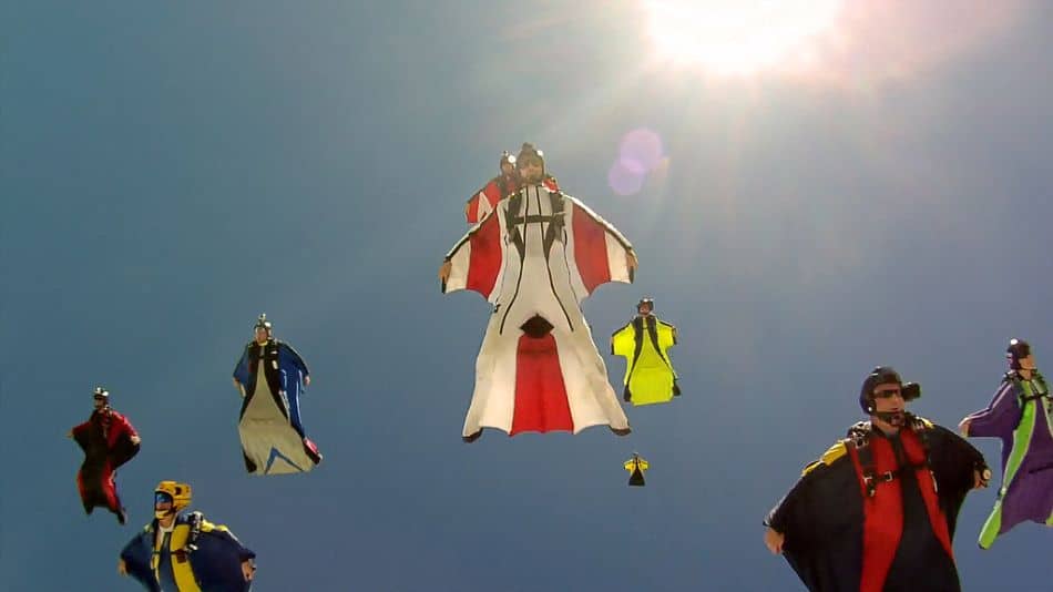 A group of eight wingsuit pilots in colorful wingsuits are captured mid-flight against a clear blue sky, with the sun shining brightly.