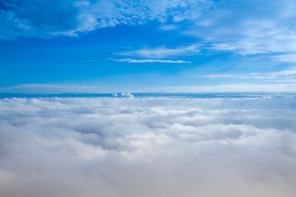 A serene view of a vast sky, where the lower half is dominated by fluffy white clouds and the upper half reveals a clear blue sky with scattered, wispy clouds.