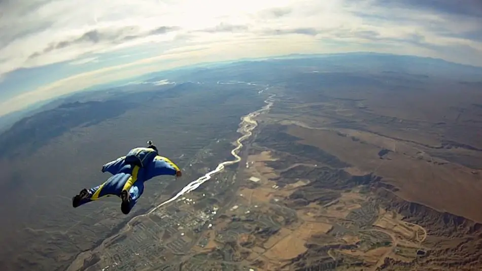 A wingsuit pilot in a blue and yellow suit captured mid-air, soaring above a vast landscape featuring a winding river, varied terrains, and scattered clouds casting shadows below. The sky is bright blue with feathery clouds.