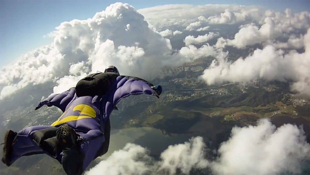 A wingsuit pilot in a blue and yellow wingsuit is soaring above the clouds, with a panoramic view of the landscape below.