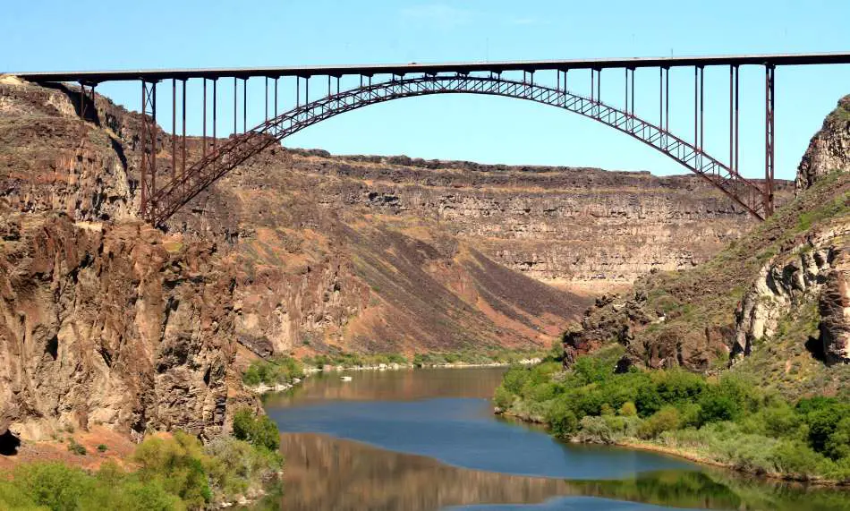 A majestic steel arch bridge spans across a serene river, nestled within a rugged canyon adorned with patches of greenery under the clear blue sky.